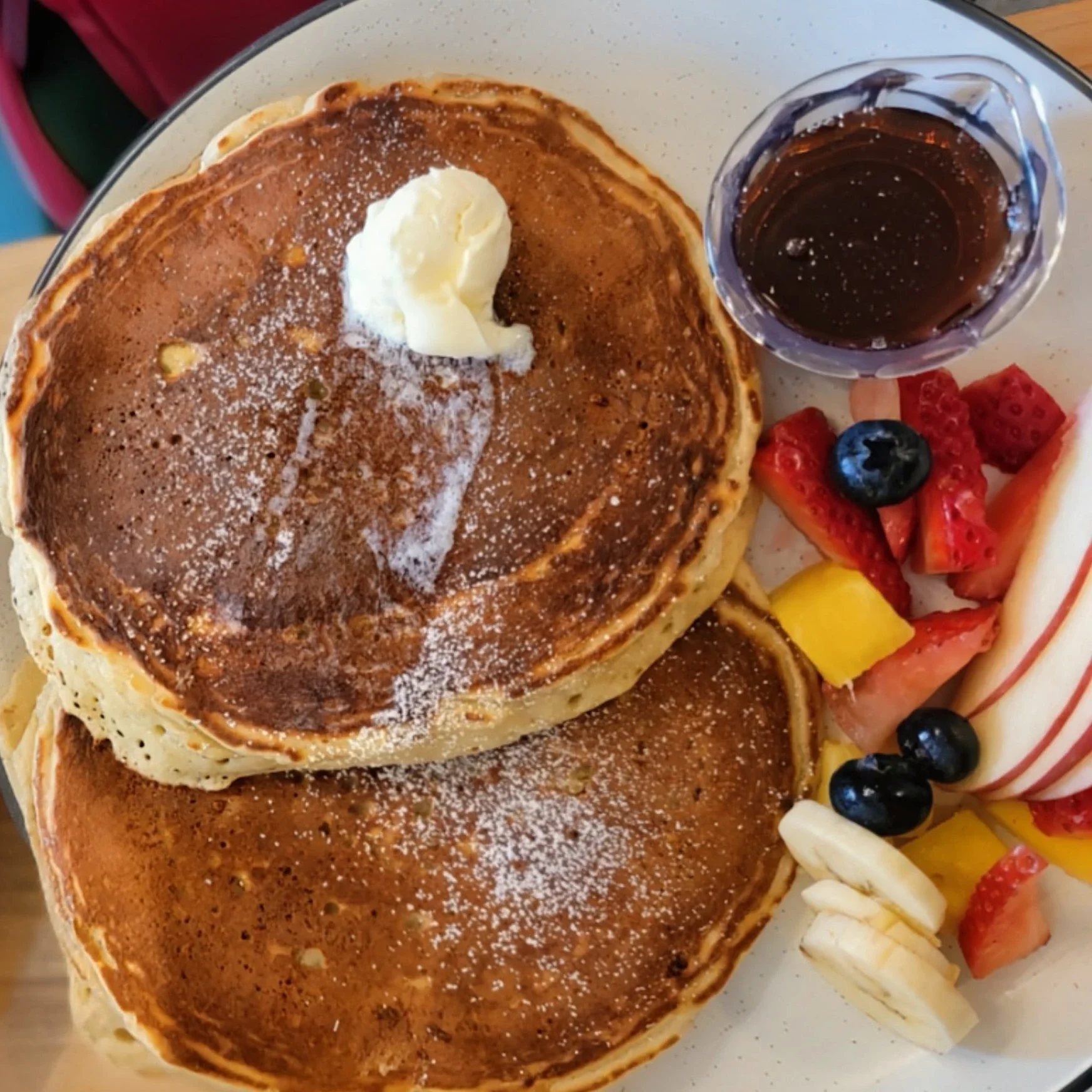 Large golden buttermilk pancake topped with melting butter and served with a side of maple syrup and fruits